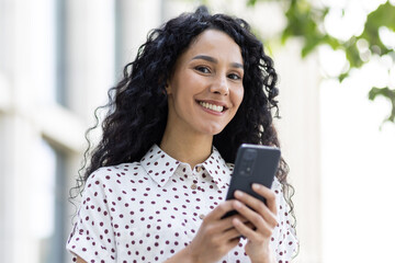 Portrait of an attractive happy young woman holding a phone, standing on the street, smiling and looking at the camera.