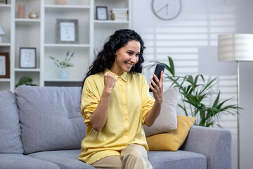 Obraz premium Happy hispanic girl sitting on sofa in living room with phone in hands, happy with good news, looking at phone screen, winner.