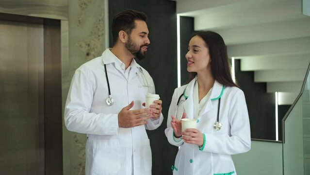 Portrait of two confident medical workers dressed in physician robe having pleasant conversation with cup of hot drink in hands. Skilled man and woman spending time together in hospital atmosphere.