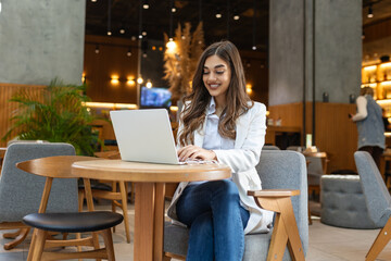 Young woman, digital nomad working remotely from a cafe, drinking coffee and using laptop, smiling.