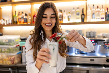 Happy young female barkeeper in stylish outfit serving winter mojito cocktail with berries while...