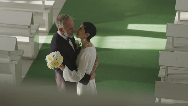 High angle slowmo of mature newlywed Caucasian couple kissing, standing on green carpet in cozy white wedding chapel after ceremony