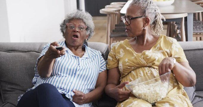 Happy senior african american female friends on couch eating popcorn, using tv remote, slow motion