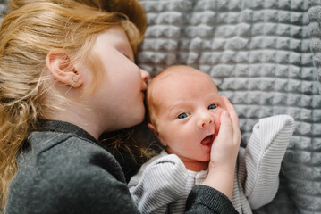 Sibling relationship in family when youngest was born. First meeting baby and toddler older sister. Young girl tenderly hugs her newborn while lying on bed at home. Children hugging together. Top view