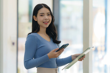 Young beautiful Asian business woman holding a folder and a smart phone while standing at the...