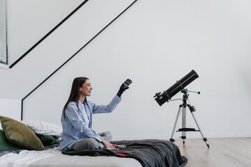 Enthusiastic woman with prosthetic arm reaching towards a telescope in a well-lit bedroom. Girl...