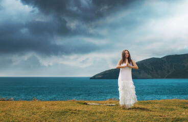 Fototapeta na wymiar A young woman, dressed in a white dress, does meditation exercises (yoga) in nature, in a beautiful landscape. Woman with hands together, as in a prayer