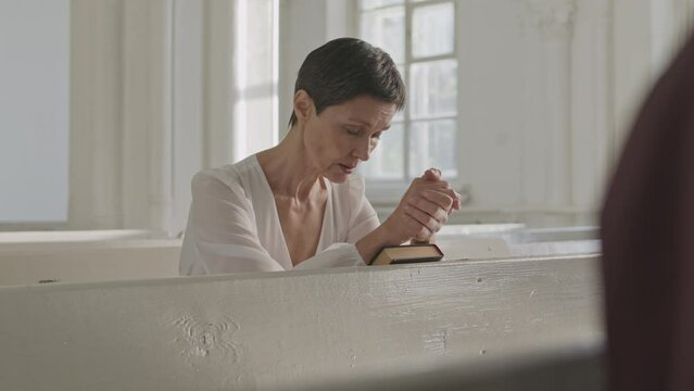 Slowmo of short haired mid-aged Caucasian woman looking down and saying prayer while sitting on white wooden bench during church service