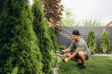 A young man in a straw hat and hands in gloves is trimming bushes in his garden with a big...
