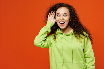 Young curious nosy fun woman of African American ethnicity she wear green hoody casual clothes try to hear you overhear listening intently isolated on plain red orange background. Lifestyle concept. - obrazy, fototapety, plakaty