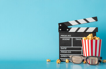 Experience excitement of much-awaited movie debut. Side view captures table scene with clapperboard, striped popcorn box, and 3D glasses against light blue wall, leaving space for text or film adverts