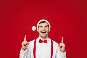 Merry young smiling cool man wear white shirt Santa hat posing point index finger overhead on area...