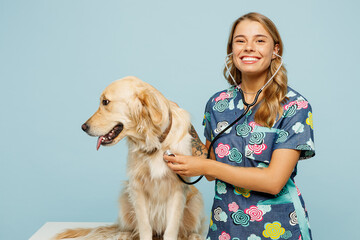 Young smiling happy veterinarian woman she wearing uniform heal exam retriever dog use stethoscope look camera isolated on plain pastel light blue background studio portrait. Pet health care concept.