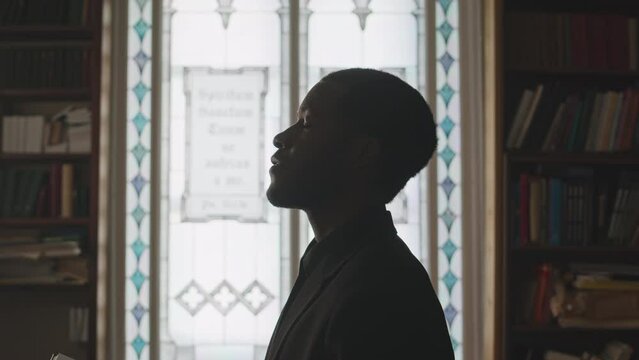 Side view tilt up slowmo of young African American pastor standing by stained glass window in church library with Bible, praying and raising his head up to God