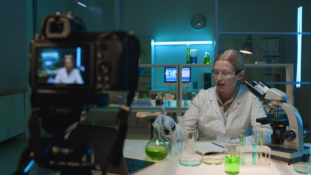 Medium selective focus shot of camera on tripod and middle-aged Caucasian female chemist sitting in lab, giving online lesson, showing flasks with solution, filming video content for educational blog