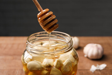 Natural honey dripping from dipper into glass jar with garlic on table, closeup
