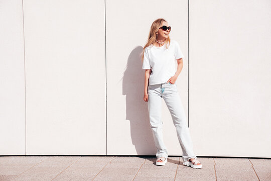 Portrait of beautiful smiling model in sunglasses. Female dressed in summer hipster white T-shirt and jeans. Posing near wall in the street. Funny and positive woman having fun outdoors