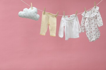 Different baby clothes and cloud shaped pillow drying on laundry line against pink background. Space for text