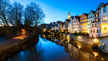Panoramic view of illuminated historic facades of old town of Tuebingen on Neckar River in southern Germany. Winter evening twilight with colorful reflections, Hölderlin tower and church. Blue hour. - obrazy, fototapety, plakaty