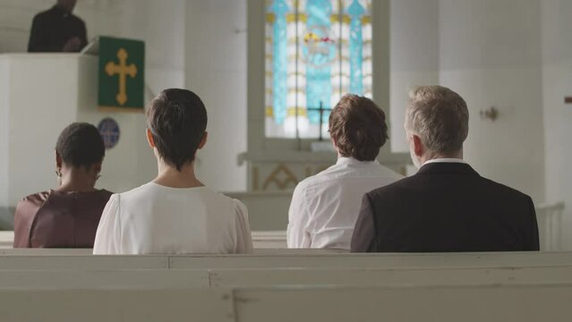 Back view slowmo of group of diverse parishioners sitting on benches in front of Black male pastor preaching sermon from tribune in Lutheran church