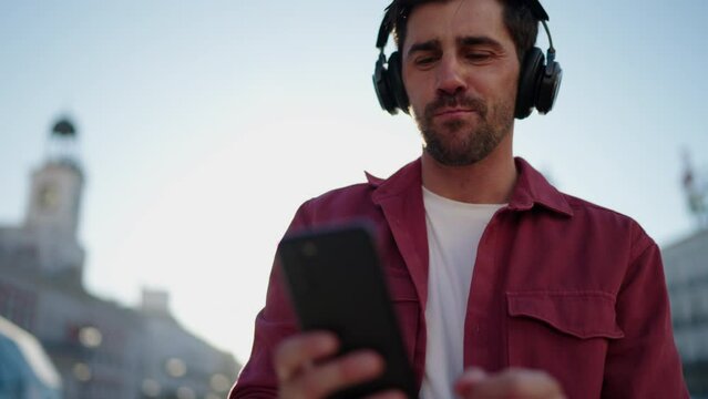 Excited man in headphones using music streaming app to find perfect song, exploring latest 6G internet capabilities for faster streaming.Happy male singing along to favorite tunes while wandering city