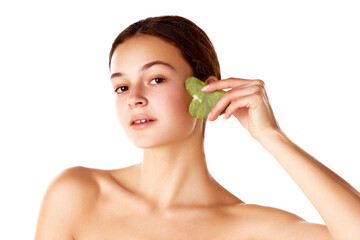 Beautiful young girl taking care after skin, using face massage tool gua sha against white background. Face lifting effect. Concept of natural female beauty, skin care, cosmetology and cosmetics