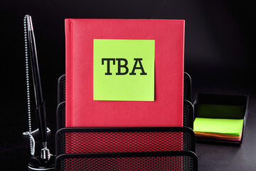 TBA Text on a yellow sticker on a red vertical notepad on a black background next to a pen and money