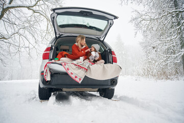 On the towel, with warm drink. Woman with her daughter is on the trunk of a car in the winter meadow and forest
