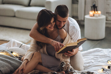 Happy young couple relaxing together in the bedroom at home, reading book. Man and woman enjoying lazy cozy weekend at home, embracing, kissing, cuddling. Simple pleasures, domestic life - obrazy, fototapety, plakaty
