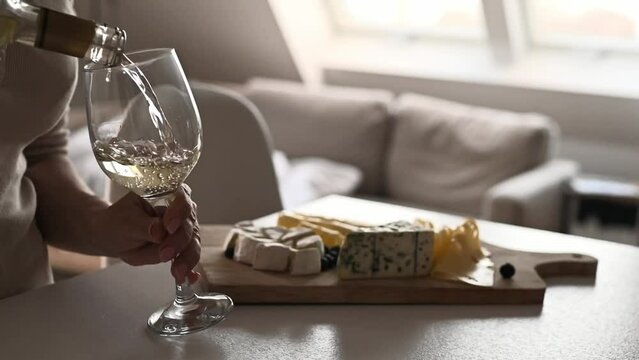 Man pouring white wine to glasses from bottle at kitchen with cheese plate on table. Transparent wineglasses and sommelier filling them with alcohol beverage