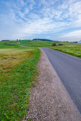 Morning vista, A winding road guides through green meadows, adorned with charming huts, beneath a partly cloudy sky and the serene embrace of blue