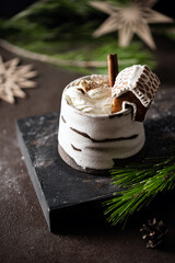 Hot winter drink in a clay mug. Close-up of hot chocolate with cinnamon stick, gingerbread house whipped cream. Winter cozy still life with cocoa, Christmas decorations, fir branch and lights. © Lizaveta
