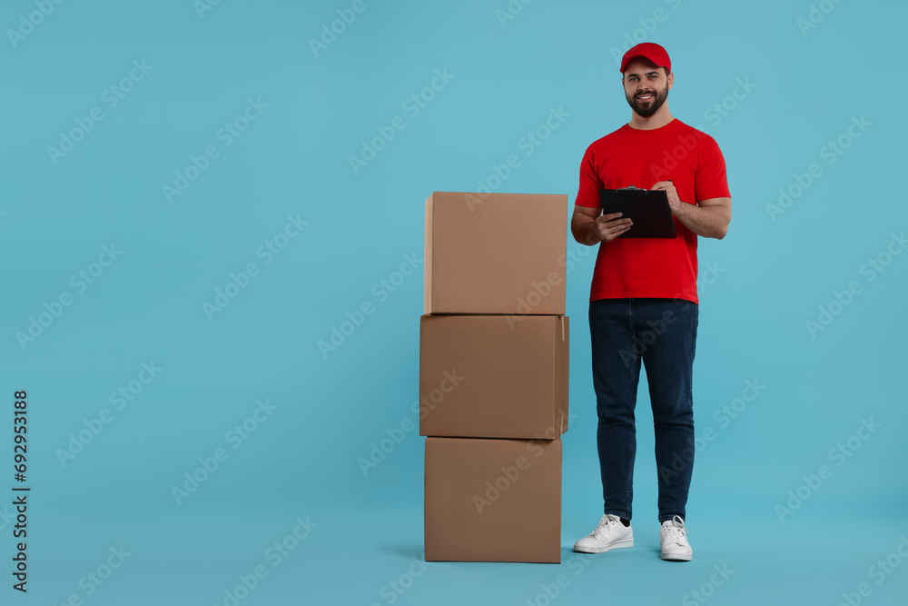 Wall mural Happy young courier with clipboard and stack of parcels on light blue background, space for text