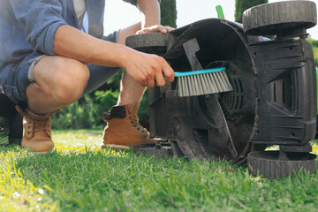 Young man cleaning lawn mower with brush in garden, closeup