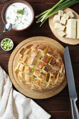 Freshly baked bread with tofu cheese, green onions, sauce and knife on wooden table, flat lay