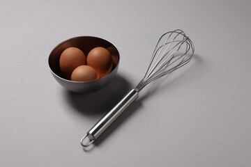 Metal whisk and raw eggs on grey background