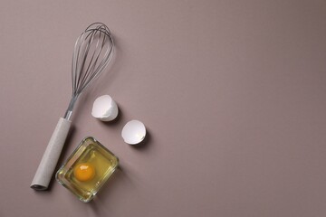 Metal whisk, raw egg in bowl and shells on grey background, flat lay. Space for text