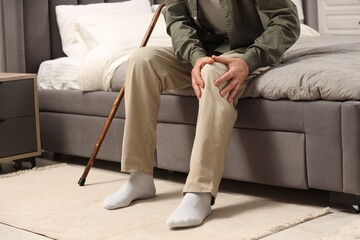 Senior man with walking cane suffering from knee pain on bed at home, closeup. Rheumatism symptom