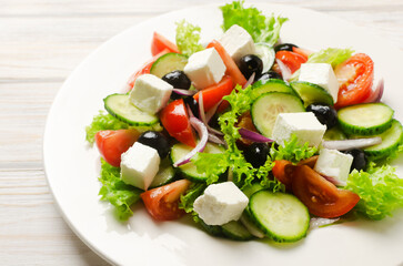 Classic Greek salad with cucumbers, tomatoes, red onion, feta cheese, lettuce and black olives on a white wooden table. The concept of traditional dishes. Healthy food. Simple ingredients.