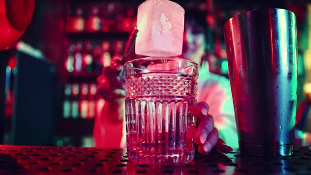 Focus on cocktail glass. Young girl, bartender working in bar, nightclub, making sweet and sour cocktail, putting ice into glass. Concept of nightlife, bar, party, alcohol drink, mixologist