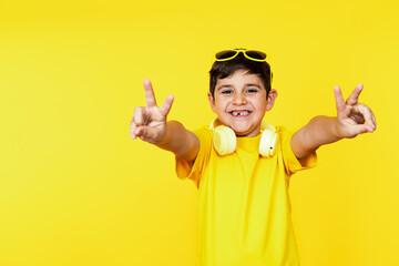 Happy Caucasian kid in yellow tee and headphones makes victory signs, vibrant yellow backdrop