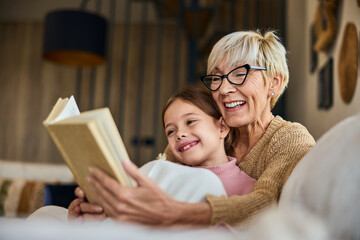 A smiling grandchild spending time with her grandma, reading a book together, covered in a blanket.