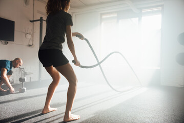 Unrecognizable sportswoman exercising with ropes in frotn of sunlit gym window