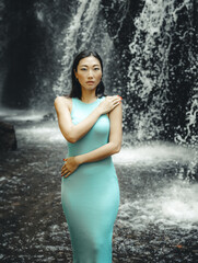 Attractive Asian woman posing near the waterfall. Water splashing. Nature and environment. Travel lifestyle. Slim woman wearing light blue dress. Copy space. Yeh Bulan waterfall in Bali
