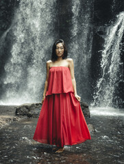 Charming Asian woman posing near waterfall. Nature and environment. Water splashing. Travel lifestyle. Young woman wearing long red dress. Slim body. Copy space. Yeh Bulan waterfall, Bali
