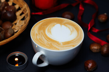 White cup of coffee, surrounded by chestnuts and red ribbons, during winter and holiday season, bright and dark contrast