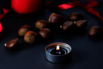 Perfumed candle burning, surrounded by chestnuts and red ribbons, on a dark background, during...