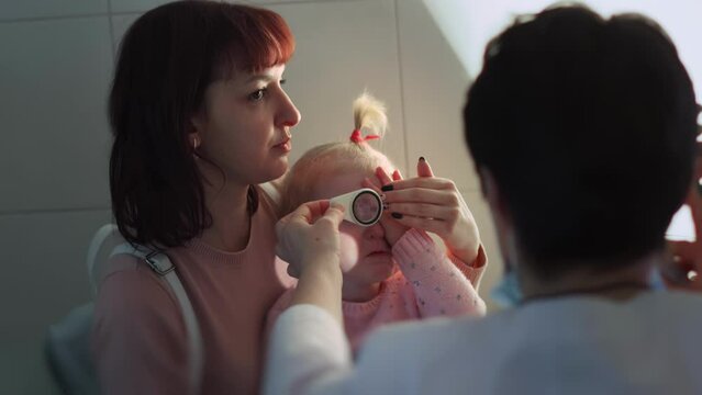 Pediatric ophthalmologist optometrist is trying to look at eyes of naughty child who cries. Doc with magnifying glass checks vision of female kid. Eye disease. Two years old little girl with mother