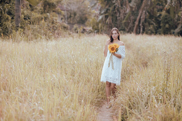 Caucasian woman walking in a field and holding a bouquet of sunflowers. Beautiful  woman wearing white dress. Summer time. Horizontal layout. Lifestyle concept.