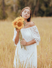 Caucasian woman walking in a field and holding a bouquet of sunflowers. Portrait of smiling woman wearing white dress. Summer vacation. Lifestyle concept. Romantic mood.